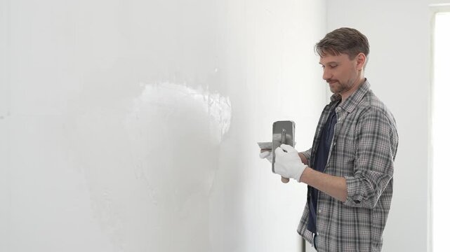 Middle aged man construction worker wearing gloves and plaid shirt is using finishing trowel to plaster white wall, applying a layer of plaster to create a smooth surface during renovation project