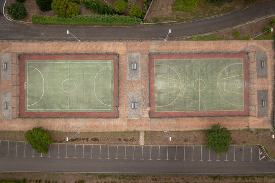 Aerial view of the sports courts at Parque Infantil De Oza featuring a field hockey pitch and a basketball court surrounded by trees and parking in Castres, A Coruna, Spain.