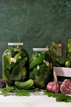 Cucumbers in a basket with dill, garlic, pickling leaves, and pickled cucumbers in jars on a kitchen counter. Harvest concept. Homemade pickle recipe and ingredients. Home canning concept.