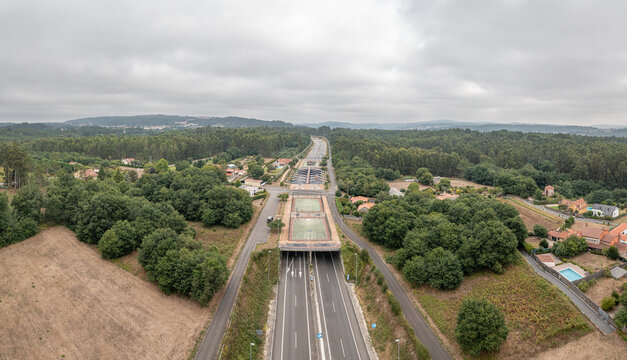 Aerial view of a highway tunnel entrance surrounded by lush green forests and rural houses under a cloudy sky in Castres, A Coruna, Spain.