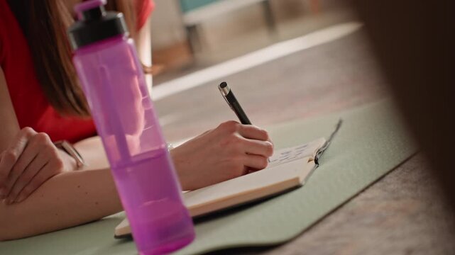 White woman at home writing workout plan on yoga mat with purple bottle, pen and notebook, cozy living room sunlight, focused wellness planning, hydration reminders and gentle calm mood
