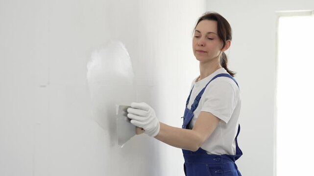 Construction worker woman spreading finishing putty on white wall, wearing protective gloves while smoothing surface with putty knife for precise home repair