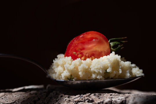 A close-up of a spoon with cooked couscous and half a tomato on it. The background is dark.