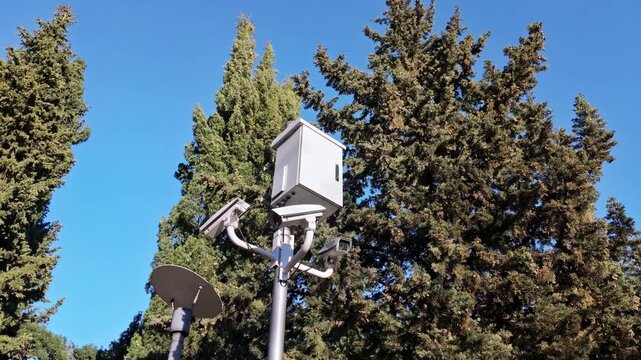 Modern urban pole equipped with security cameras and light fixture stands among green trees in park.