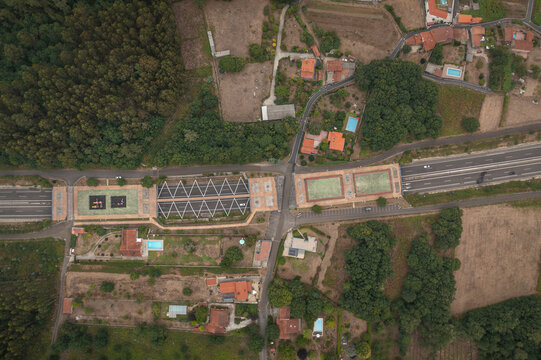Aerial view of Parque Infantil De Oza featuring sports courts and recreational areas built over a highway surrounded by lush greenery and rural houses in Castres, A Coruna, Spain.