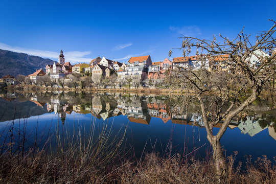 View of the historic town reflected in the calm waters of the Mur river under a clear blue sky in Frohnleiten, Styria, Austria.