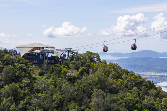 View of the Langkawi SkyCab cable car station on a lush mountain peak with red gondolas traveling along cables against a backdrop of blue sky in Langkawi, Kedah, Malaysia.