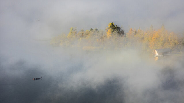 View of Hallstatter See shrouded in morning fog with autumn trees, a small wooden boat, and a cabin on the calm water in Salzkammergut, Upper Austria, Austria.