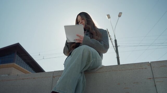 Asian woman on ledge holding journal in sunlight. Casual dressed woman gazes warmly with journal on city ledge. Sunshine illuminates optimistic female figure with notebook atop city balcony ledge