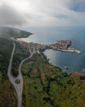 Aerial view of the coastal town of Caion with its harbour, rocky coastline, and winding road surrounded by green hills in Caion, A Coruna, Spain.