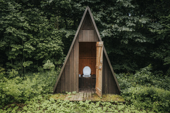 Wooden A-frame outdoor toilet structure with an open door, surrounded by lush greenery and dense foliage in a forested area