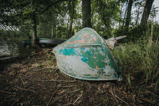 Abandoned weathered boat resting on the shore of a lake surrounded by trees and overgrown grass, showcasing peeling paint and rustic textures