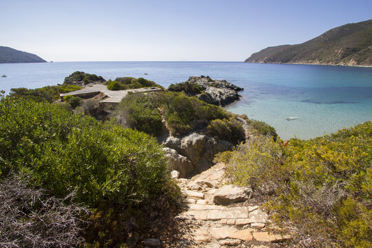 View of the rocky path leading down to the turquoise waters of Spiaggia Laconella, surrounded by Mediterranean vegetation under a clear blue sky in Elba, Toscana, Italy.