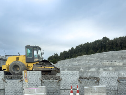 View of a yellow road roller operating at a construction site with concrete retaining walls and a forested hillside in the background in Sariyer, Istanbul, Turkiye.