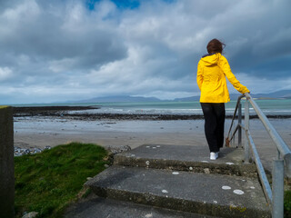 Person in bright yellow jacket stands on stone steps by coastal path. They face calm sea under cloudy sky. View shows distant shoreline and rocky beach © mark_gusev
