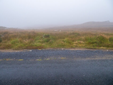 Foggy narrow road stretches into misty distance. Grassy fields flank narrow asphalt path. Mist hides horizon and surrounding terrain. No visible animals or specific plants. Dangerous driving area