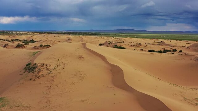 Aerial view of Gobi Desert sand dunes with sparse vegetation and distant green plains under a dramatic cloudy sky in Nomgon, Omnogovi, Mongolia.
