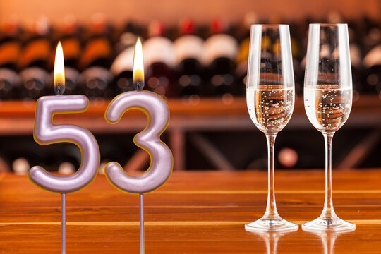 Birthday card, anniversary or celebration of a special day, lit candles forming the number 53 and two champagne glasses; Photo on a background of a bookshelf with blurred wine bottles.