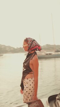 Elegant woman wearing a headscarf and sunglasses walking along the waterfront past a classic convertible car during a sunny day, embodying a chic and luxurious summer vacation lifestyle