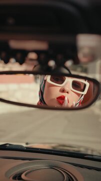 Mysterious beautiful woman with sunglasses and red lipstick looking at her reflection in the rearview mirror while sitting inside a vintage car, embodying a retro and cinematic style. Vertical video