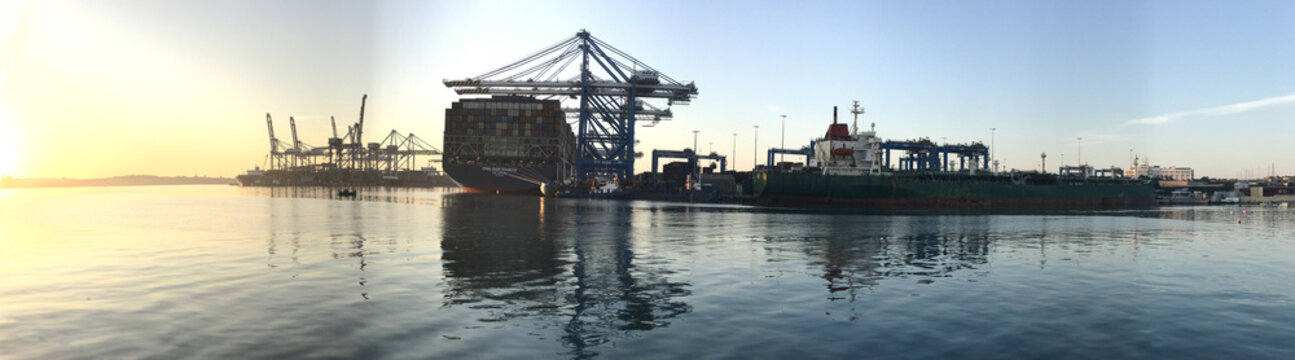 View of Malta Freeport Corporation Ltd with large container ships and gantry cranes reflecting on the calm water at sunset in Birzebbuga, Southern Region, Malta.