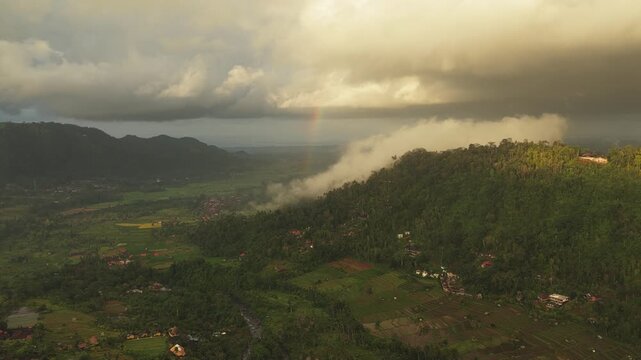 Aerial view of Sidemen Rice Terrace with lush green hills, misty clouds, and a faint rainbow over the valley in Kecamatan Sidemen, Bali, Indonesia.