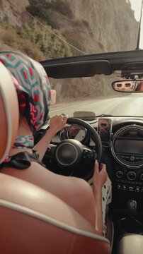 Beautiful woman with headscarf driving a cabriolet on a scenic mountain road during a summer road trip, enjoying the journey and the feeling of freedom on a sunny day along the coast. Vertical video