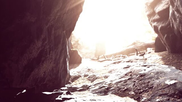 Visitors walk through a rocky cave with sunlight coming in from the entrance. The cave has rough stone surfaces and a natural path that leads outside.