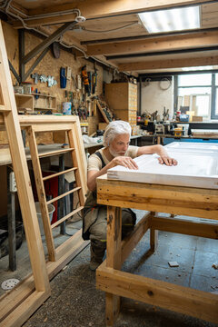 Attentive carpenter examines antique wooden door at worktable in restoration woodworking workshop, man checking damage of paint, assesses quality of wood, crafter at workbench inspects old door.