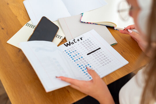 A high-angle close-up shows a girl holding an open journal with a written water tracker at a desk. Multiple notebooks and a smartphone rest on the table as she writes.