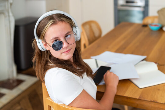 A teenage girl wearing eyeglasses with a corrective eye patch for strabismus treatment looks at the camera while doing homework indoors. Headphones rest on her head as she studies at a wooden table.