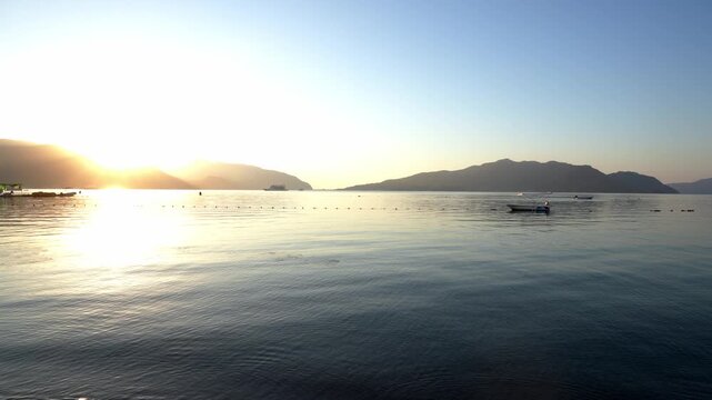 Scenic waterfront in Marmaris, Turkey during blue hour at sunrise or sunset. The coastal promenade overlooks calm sea framed by mountain silhouettes in the distance. Small boat floats gently on waves