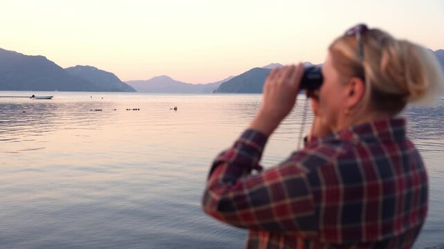 Defocused Woman standing on the waterfront in Marmaris, Turkey looks through binoculars toward the sea during golden hour at sunrise or sunset. Coastal promenade overlooks calm water and distant