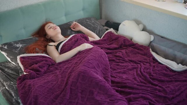 Young caucasian woman gazing at ceiling in bed under purple blanket, relaxed hands and dreamy expression, morning light through window, decorative shelf nearby, atmosphere of gentle anticipation
