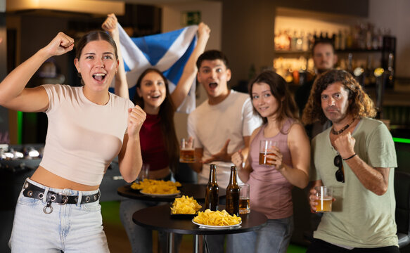 Cheerful football fans waving the flag of Scotland while drinking beer and watching tournament in sport bar