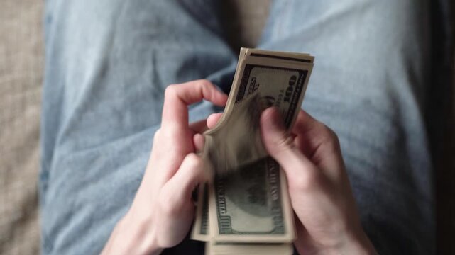 Money. A man counts a stack of hundred dollar bills in his hands. The man continues to manipulate the cash, focusing on the detailed texture of the bills.