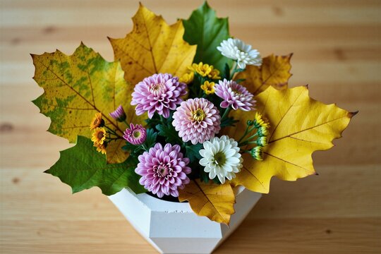 Geometric planter holds autumn flowers and yellow maple leaves arrangement. Chrysanthemums bloom with small yellow and white florets. Natural fall decor on wooden table.