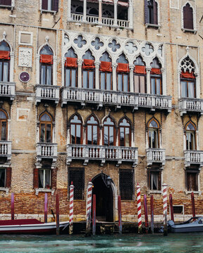View of a historic Venetian Gothic palace with intricate pointed arches, stone balconies, and red curtains overlooking a canal with traditional striped mooring poles in Venice, Veneto, Italy.