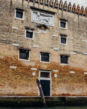 View of the historic brick facade featuring the Lion of Saint Mark relief, barred windows, and a dark wooden door along a canal in Venice, Veneto, Italy.