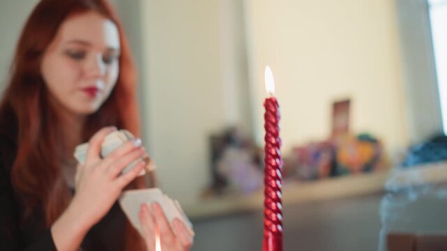 White redhead shuffles tarot cards near burning candle, hands and deck in frame with flickering flame in foreground, focused fortune teller gesture creates tense, expectant mood amid colorful altar