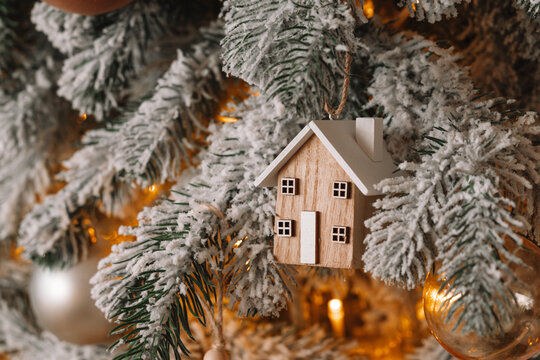 Cozy Christmas tree snow covered branch decorated with wooden house ornament, warm lights garland and golden baubles, creating festive winter atmosphere. Festive aesthetic atmosphere, holiday mood