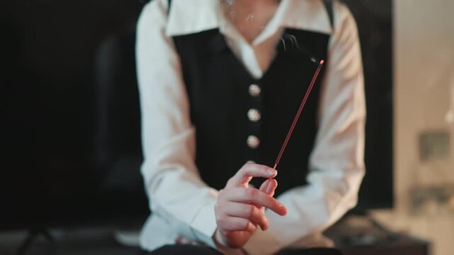 Woman holding burning incense stick in intimate indoor reading scene, black vest white blouse, delicate hands and fingers, smoldering smoke trails rising, soft candlelight glow, contemplative mood,