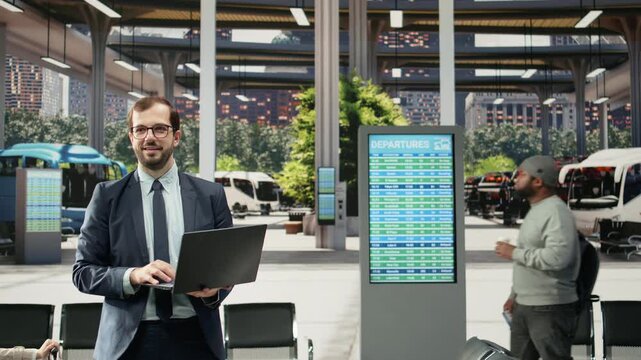 Happy business traveler stands in a bus station with a notebook while waiting for departure, holding a laptop and preparing for a coach connection during a workday commute and business journey.