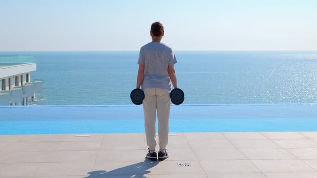 Woman walking with dumbbells near infinity pool and sea. Woman performing a farmer's walk exercise with dumbbells along the edge of an infinity pool, with a sea view in the background on a bright