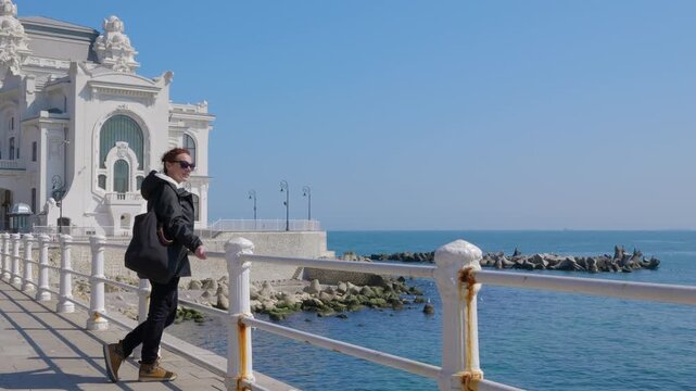 Female tourist walking on seaside promenade admiring the constanta casino. Young woman wearing a backpack walking on a seaside boardwalk on a sunny day. Stopping to admire the view of the black sea