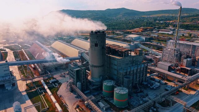 White steam rises from various vents across a huge manufacturing complex. Large industrial plant against green hills.