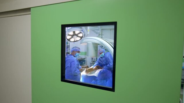 Green door with the square window to the modern surgery. Doctors work inside the rom performing operation.