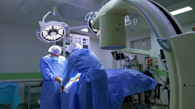 Doctors in blue uniforms stand at the operating table covered with blue sheet. Two round lamps are directed on the surgeons performing surgery on foot.