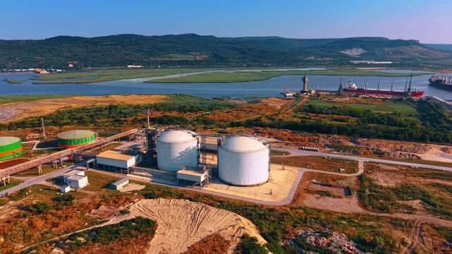 Heading to two massive white spherical pressurized tanks near an open river delta surrounded by green hills. Some bulk carrier vessels in the cargo terminal dock in the backdrop.