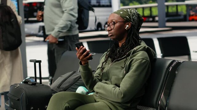 Woman sends voice notes on a phone in a bus station while wearing earbuds during travel, recording a message on her smartphone before departure.